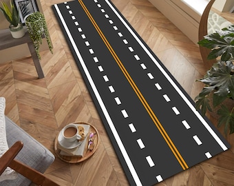 以下が含まれることがあります： 道路のデザインが施された長方形の黒いラグ。ラグは、高速道路を模倣した、側面に白い破線と中央に黄色の二重線が特徴です。 ラグは木製の床に置かれています。