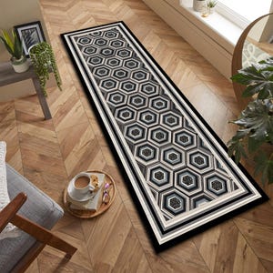May include: A long, rectangular rug with a geometric honeycomb pattern in shades of black, gray, and white. The rug has a black border with a white and gray inner border. The rug is placed on a wooden floor.