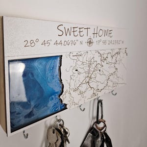 May include: A wooden key holder with a map design and the words "SWEET HOME". The key holder features a blue resin section and a map outline. It includes several hooks for keys and a compass symbol.