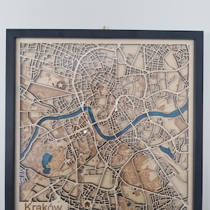 Könnte beinhalten: Eine gerahmte Holzkarte von Krakau, Polen, mit einem schwarzen Rahmen. Die Karte ist aufwendig geschnitten und zeigt Straßen, Gebäude und einen blauen Fluss. Das Wort "Kraków" befindet sich unten links.