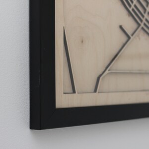 May include: Close-up of a framed wooden map with a black frame. The map is light-coloured with dark lines depicting streets and other features. The frame is black and the background is a white wall.