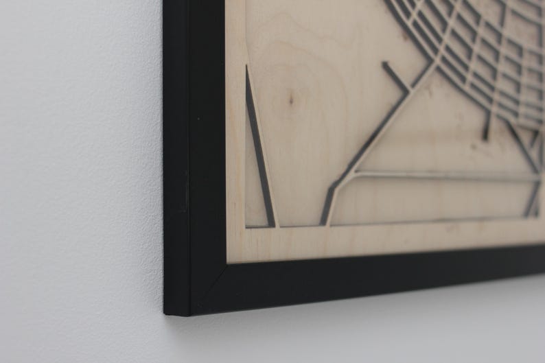 May include: Close-up of a framed wooden map with a black frame. The map is light-coloured with dark lines depicting streets and other features. The frame is black and the background is a white wall.