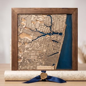 Könnte beinhalten: Ein quadratisches Wandbild mit Holzrahmen, das eine lasergeschnittene, mehrschichtige Karte von Asbury Park, NJ zeigt. Die Karte zeigt komplexe Straßennetze in hellen Holztönen, kontrastiert durch einen tiefblauen Harzabschnitt.