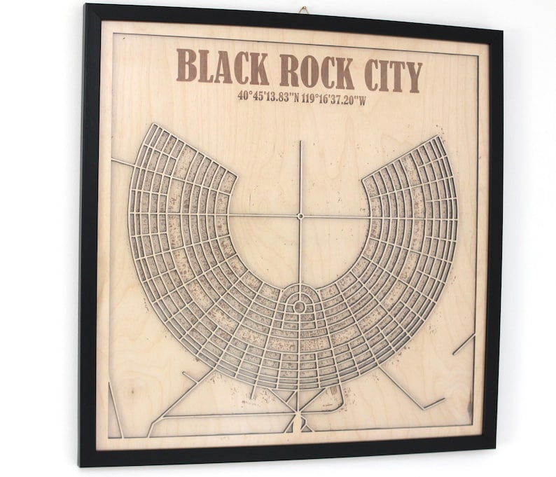 May include: A framed wooden map of Black Rock City, with the city's name and coordinates printed at the top. The map features a detailed, radial design of the city's layout, with a black frame. The wood has a light, natural colour.