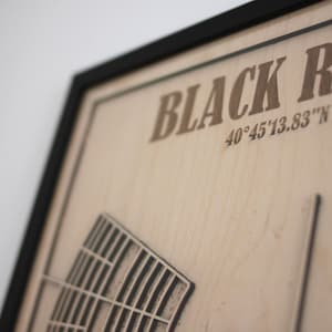 May include: Wooden framed artwork with the words "BLACK ROCK" and coordinates 40&deg;45'13.83"N 119&deg;16'37". The artwork features a detailed, carved design of a structure. The frame is black.