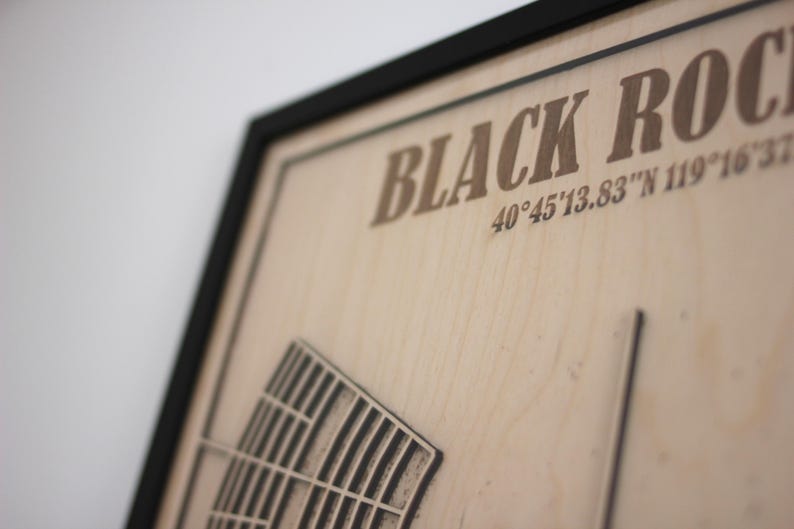 May include: Wooden framed artwork with the words "BLACK ROCK" and coordinates 40&deg;45'13.83"N 119&deg;16'37". The artwork features a detailed, carved design of a structure. The frame is black.