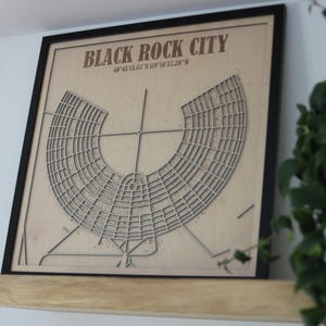 May include: A framed wooden map of Black Rock City, with the city's name and coordinates in brown text. The map has a detailed, geometric design within a black frame, suitable for home decor.