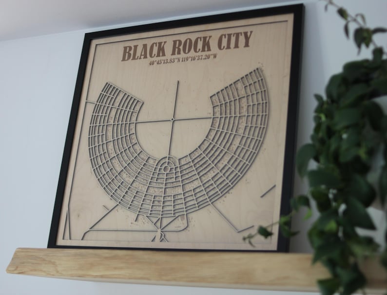 May include: A framed wooden map of Black Rock City, with the city's name and coordinates in brown text. The map has a detailed, geometric design within a black frame, suitable for home decor.