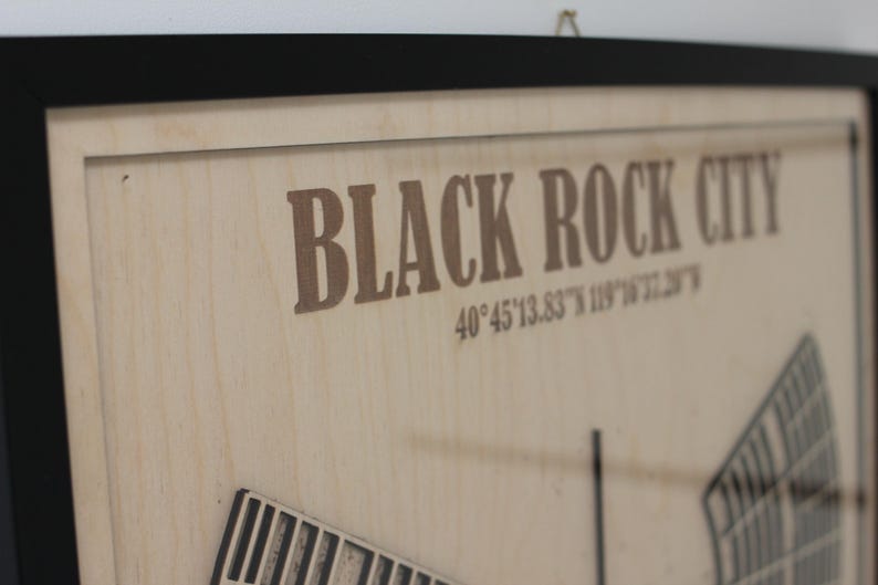 May include: Wooden framed artwork featuring the words "BLACK ROCK CITY" in brown lettering. The artwork also includes coordinates and a partial architectural rendering. The frame is black.