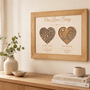 May include: A framed wall art piece featuring the text Our Love Story with two heart-shaped cutouts containing detailed wooden map engravings. Below the hearts, the text reads FIRST MET October 2015 London and ENGAGED 10.01.2022 Zakopane.