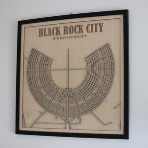 May include: A framed wooden map of Black Rock City, Nevada, with the city's layout etched into the wood. The map is in a black frame and includes the city's coordinates. The text "BLACK ROCK CITY" is at the top.