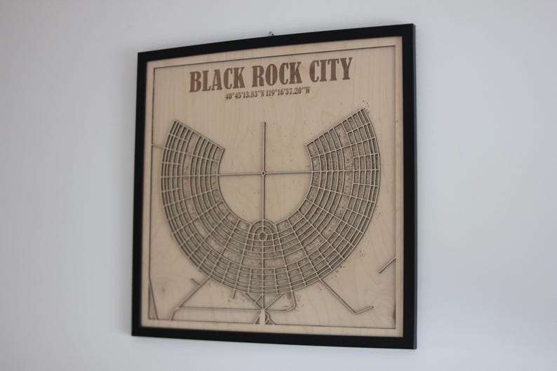 May include: A framed wooden map of Black Rock City, Nevada, with the city's layout etched into the wood. The map is in a black frame and includes the city's coordinates. The text "BLACK ROCK CITY" is at the top.