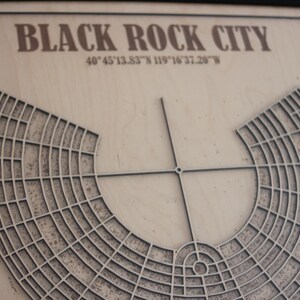May include: A framed wooden map of Black Rock City, with the city's name and coordinates at the top. The map features a detailed, layered design of the city's layout, with a circular central area and radial streets. The frame is black.