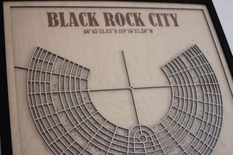 May include: A framed wooden map of Black Rock City, with the city's name and coordinates at the top. The map features a detailed, layered design of the city's layout, with a circular central area and radial streets. The frame is black.