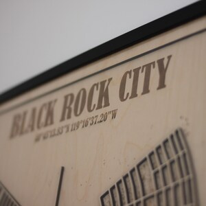 May include: A framed wooden map of Black Rock City, with the city name and coordinates printed in a brown font. The map features a detailed layout of the city's design.