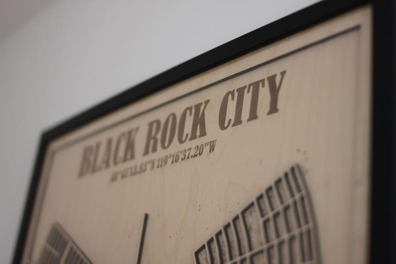 May include: A framed wooden map of Black Rock City, with the city name and coordinates printed in a brown font. The map features a detailed layout of the city's design.