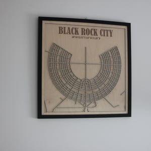 May include: Framed wooden artwork showing a laser-cut map of Black Rock City. The artwork includes the city's name and coordinates. The map is a light brown colour and is set against a black frame.