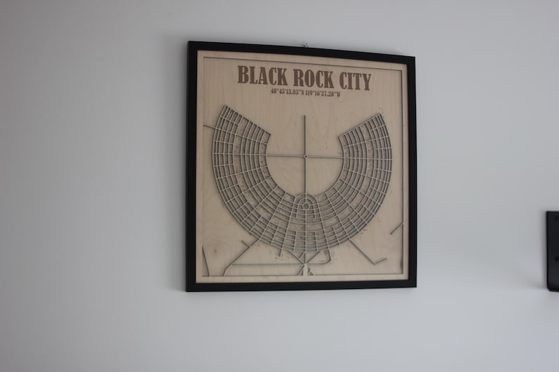 May include: Framed wooden artwork showing a laser-cut map of Black Rock City. The artwork includes the city's name and coordinates. The map is a light brown colour and is set against a black frame.