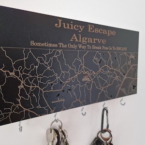 Könnte beinhalten: Schwarzer Holz-Schlüsselhalter mit einer Karte der Algarve und dem Schriftzug "Juicy Escape Algarve". Er hat Haken für Schlüssel und ein Zitat. Das Kartendesign ist detailliert.