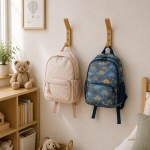 Peut inclure: Deux sacs à dos suspendus à des crochets muraux en bois. Le sac à dos de gauche est rose clair avec un motif floral subtil, une poche avant zippée et des poches latérales en filet. Celui de droite est bleu foncé avec des motifs de dinosaures.