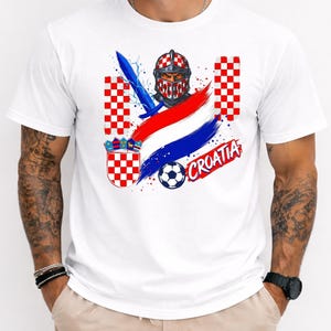 May include: A white t-shirt featuring a graphic print of a knight in a red and white checkered helmet, a blue sword, a soccer ball, the Croatian coat of arms, red, white, and blue brush strokes, and the text CROATIA in a red stylized font.
