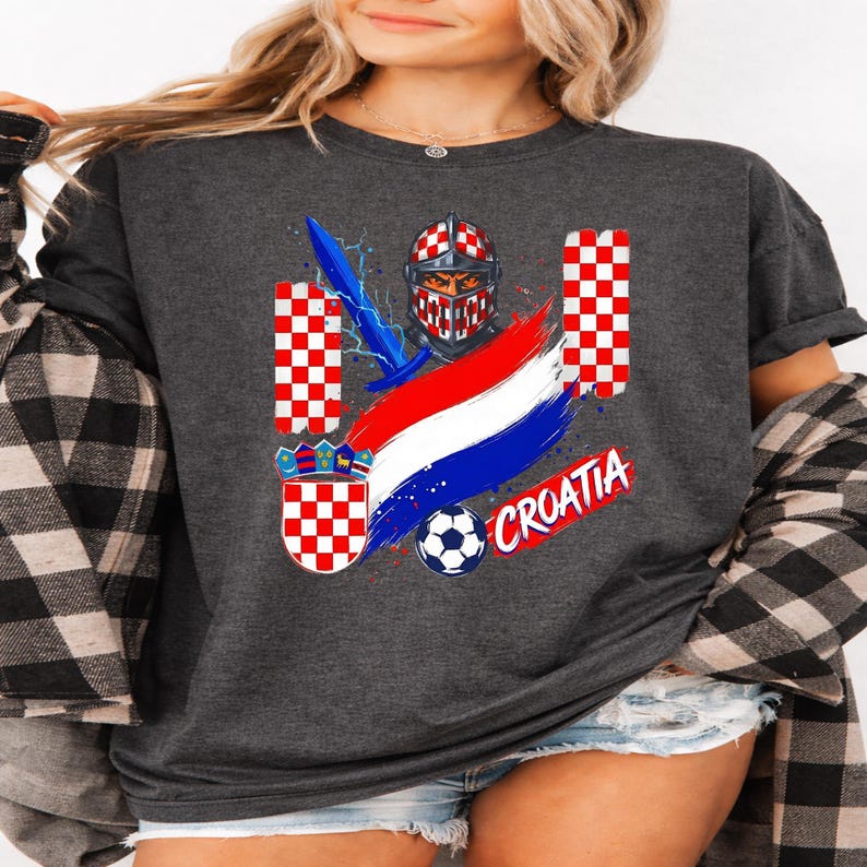 May include: A charcoal gray t-shirt featuring a graphic print of a knight in a red and white checkered helmet, a blue sword, a soccer ball, and the word CROATIA in red. The design includes red and white checkered patterns and a shield emblem.
