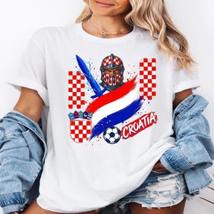 May include: A white t-shirt featuring a graphic print of a knight helmet with a red and white checkered pattern, a blue sword, a soccer ball, the Croatian coat of arms, red and white checkered columns, and the text CROATIA in red lettering.