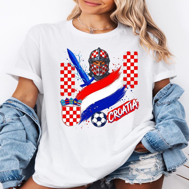 May include: A white t-shirt featuring a graphic print of a knight helmet with a red and white checkered pattern, a blue sword, a soccer ball, the Croatian coat of arms, red and white checkered columns, and the text CROATIA in red lettering.