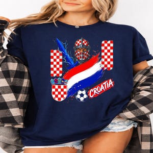 May include: A navy blue t-shirt featuring a graphic print of a knight in a red and white checkered helmet, a blue sword, a soccer ball, the word CROATIA in red, and a red and white checkered shield with a crown crest on a blue background.