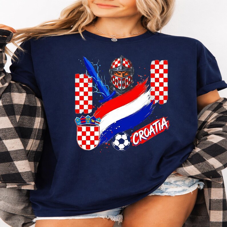 May include: A navy blue t-shirt featuring a graphic print of a knight in a red and white checkered helmet, a blue sword, a soccer ball, the word CROATIA in red, and a red and white checkered shield with a crown crest on a blue background.
