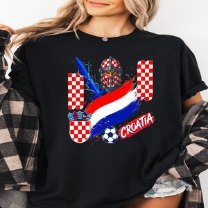 May include: A black t-shirt featuring a graphic print of a knight in a red and white checkered helmet, a blue sword, a soccer ball, and the word CROATIA in red letters. The design includes red and white checkered patterns and a tricolor flag.