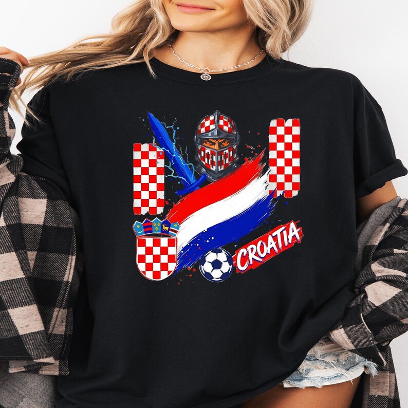 May include: A black t-shirt featuring a graphic print of a knight in a red and white checkered helmet, a blue sword, a soccer ball, and the word CROATIA in red letters. The design includes red and white checkered patterns and a tricolor flag.