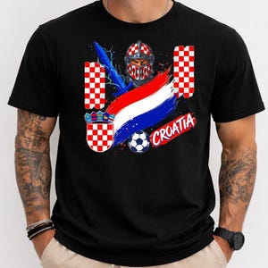 May include: A black t-shirt featuring a graphic design with a knight in a red and white checkered helmet, a blue sword, a soccer ball, the Croatian coat of arms, a red, white, and blue brushstroke flag, and the text CROATIA in red letters.