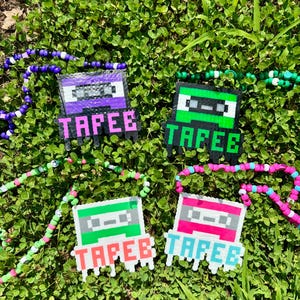 May include: Four beaded necklaces with cassette tape pendants, each with the word "TAPEE". The pendants are in purple, green, pink, and green, with matching beaded necklaces. The necklaces are on a bed of green clover.