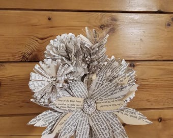 May include: A handmade paper flower bouquet. The flowers are crafted from the pages of a book, with visible text. The bouquet features a central decorative button and is set against a wooden background.