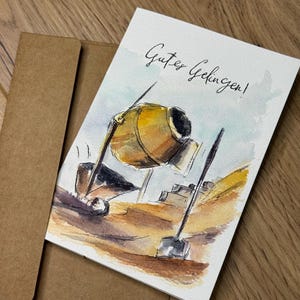 Könnte beinhalten: Aquarell-Grußkarte mit einem Baustellenthema. Die Karte zeigt einen gelben Betonmischer und den Text "Gutes Gelingen!" in schwarzer Schrift. Ein brauner Umschlag befindet sich hinter der Karte.