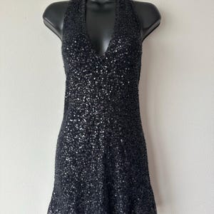 Puede incluir: Un vestido corto negro cubierto de lentejuelas, con un escote en V profundo y diseño de cuello halter. Toda la superficie de la prenda está adornada con pequeñas lentejuelas negras reflectantes que crean una textura uniforme y brillante.