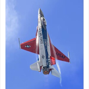 Puede incluir: Vista aérea de un avión de combate F-16 de la Fuerza Aérea de EE. UU. contra un cielo azul claro. La aeronave está pintada en rojo, blanco y azul, con "U.S. AIR FORCE" y "USAF" en un lugar destacado. El avión está en vuelo, dejando una estela de vapor.