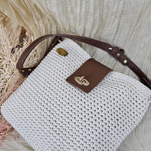 May include: A white, crocheted handbag with a brown leather strap and flap. The bag features a gold-colored clasp and a small, round gold-colored accent on the top. The strap is adjustable and has gold-colored rivets.