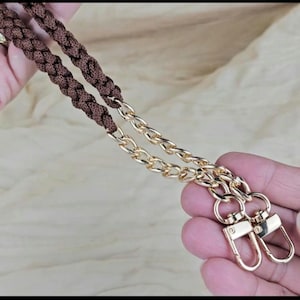 Può includere: Un cinturino intrecciato marrone con maglie a catena e chiusure color oro. Il cinturino è tenuto tra le mani di una persona, mostrando i dettagli dell'hardware e la consistenza del materiale intrecciato. Le chiusure sono pronte per essere attaccate a una borsa.
