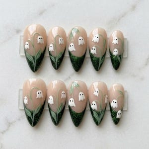 Può includere: Un set di dieci unghie finte a mandorla con base color carne. Ogni unghia presenta una punta glitterata verde scuro e steli verdi dipinti a mano con piccole figure di fantasmi bianchi che ricordano i mughetti, completi di occhi neri.