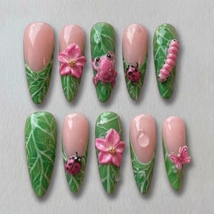 May include: A set of ten almond-shaped artificial nails. Each nail features a pink base with a white tip, and a green leaf design. Some nails have 3D embellishments of a pink flower, a frog, a ladybug, a caterpillar, and a butterfly.