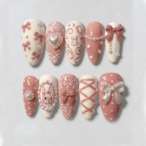 May include: A set of ten artificial nails in various designs. The nails feature a color palette of white and pink, with embellishments like bows, pearls, and a bunny illustration. Some nails have lace and heart details.