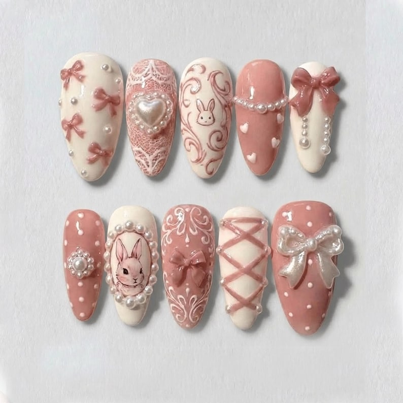 May include: A set of ten artificial nails in various designs. The nails feature a color palette of white and pink, with embellishments like bows, pearls, and a bunny illustration. Some nails have lace and heart details.