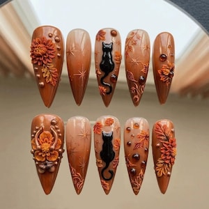 May include: A set of ten almond-shaped press-on nails featuring an autumn theme with shades of brown and orange. Designs include 3D sculpted orange flowers, black cat silhouettes, gold starbursts, amber-coloured rhinestones, and maple leaf accents.