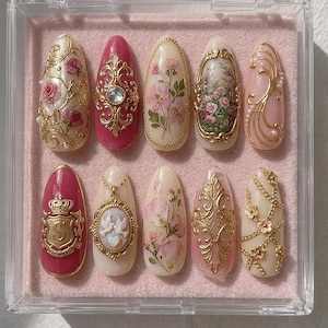 May include: A set of ten almond-shaped press-on nails arranged in a clear plastic case. The designs feature cream and deep pink bases adorned with gold filigree, floral patterns, a gold crest with the letter S, and a cameo with two white doves.