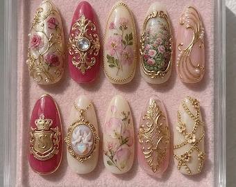 Puede incluir: Un juego de diez uñas postizas en forma de almendra dispuestas en un estuche de plástico transparente. Los diseños presentan bases en crema y rosa intenso adornadas con filigrana dorada, flores, un escudo dorado con la letra S y un camafeo.