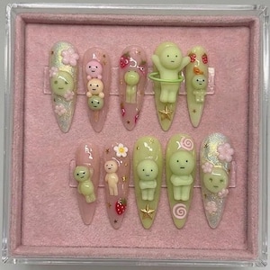 May include: A set of ten almond-shaped press-on nails featuring 3D charms of small pale green figures, pink flowers, strawberries, and gold stars. The base colors include translucent pink, solid pale green, and iridescent glitter finishes.