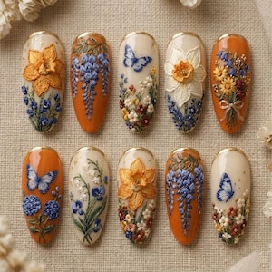 May include: A set of ten almond-shaped press-on nails featuring intricate 3D embroidery-style floral and butterfly designs. The collection includes cream and burnt orange base colors with detailed depictions of daffodils, wisteria, and blue butterflies.