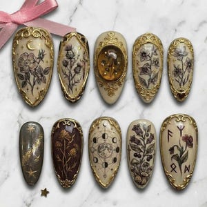 May include: A set of ten almond-shaped press-on nails with intricate designs. Each nail features a cream-colored base with gold filigree edges. Designs include floral, celestial, and botanical illustrations, with a few featuring runes and a moon phase graphic.